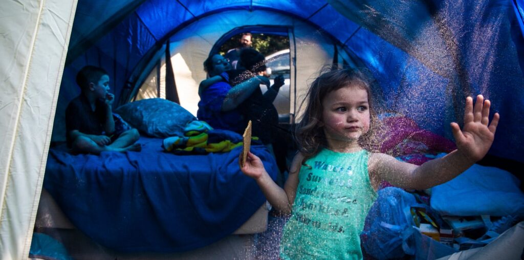 Laya Lupien eats a graham cracker and rests her hand on the door of her family’s tent while trying to escape the summer heat with her brother Dylan (left), mother Mariah, brother Evan, and father Patrick in 2019. The day before, the Lupien family of five was evicted from their home after having their Social Security income taken away, their food stamps reduced, and their rent raised without warning over the span of one year. “Thank you, mommy” said Laya for seemingly no reason. “I really don’t know why you’re thanking me,” said Mariah. “My kids are camping with no roof over their head, and they’re still thankful.”