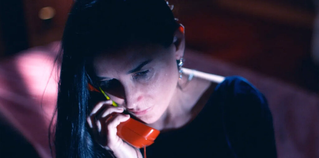 The Substance, Demi Moore's character talking into a red telephone, in a darkened room.