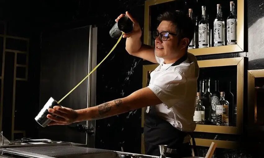 Bartender in a white shirt and black apron pours green liquid between the two cups of a metal drink shaker over an industrial bar sink. Behind him are bottles of various liquors on wooden shelving made to look like frames on a wall.