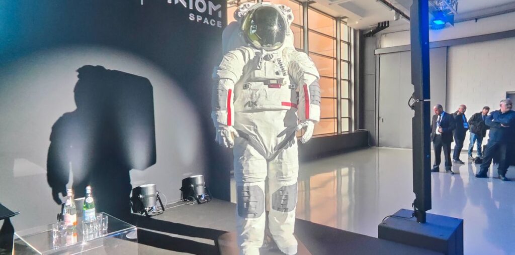 photo of a white spacesuit standing on stage at a conference, with a handful of onlookers in the background