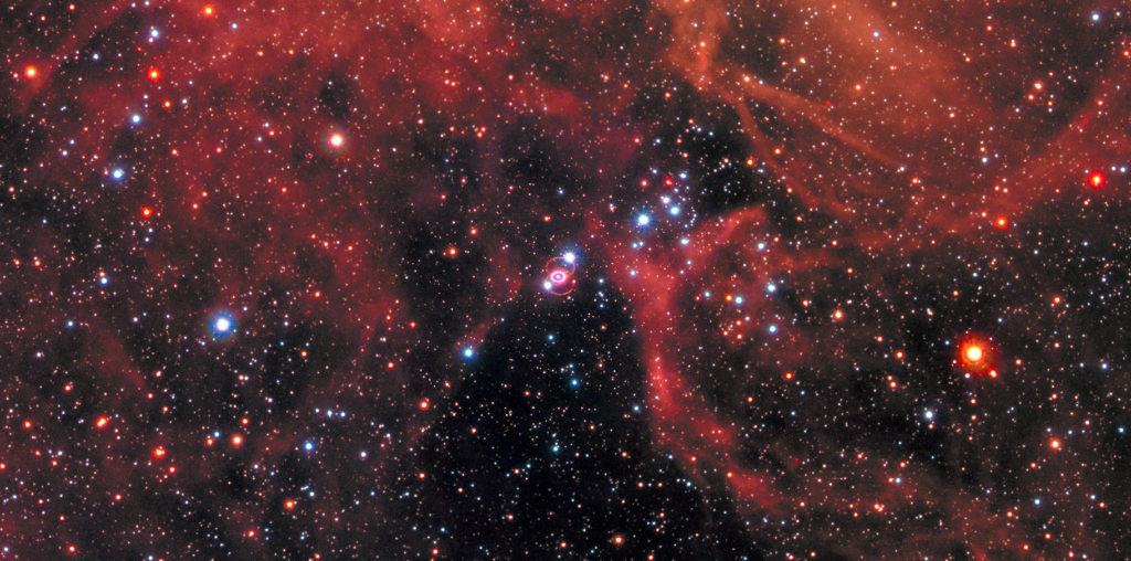 Clouds of gas and dust in shades of red cover the top of the image and a little bit of the bottom. Right in the middle is Supernova 1987a, the remnant of an exploded star. The site of the supernova is surrounded by an illuminated pinkish ring. Two faint outer rings are also visible. Stars and galaxies shine throughout, giving off red and blue light.