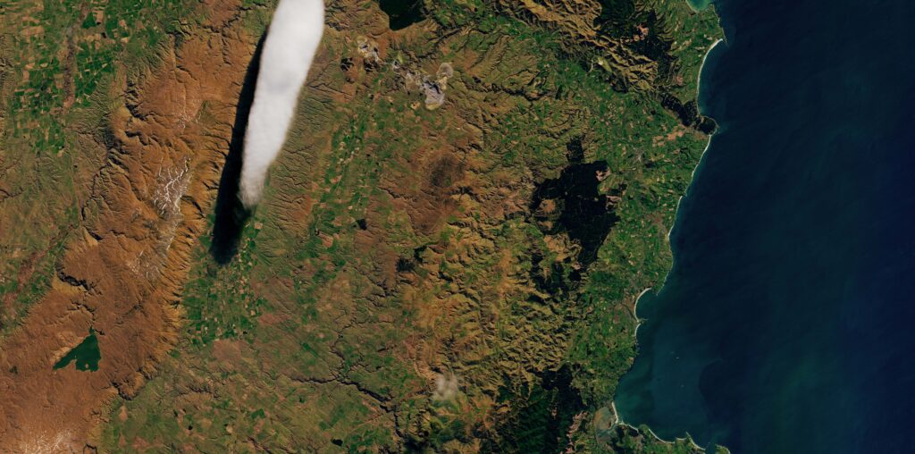 A part of New Zealand's South Island as seen by the Landsat 8 satellite. A long, thin white cloud floats above the green and brown landscape. A bit of water is visible on the right of the image.