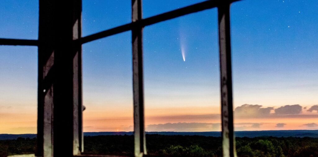 a comet streaks the sky above a dark forest seen through empty windows high up