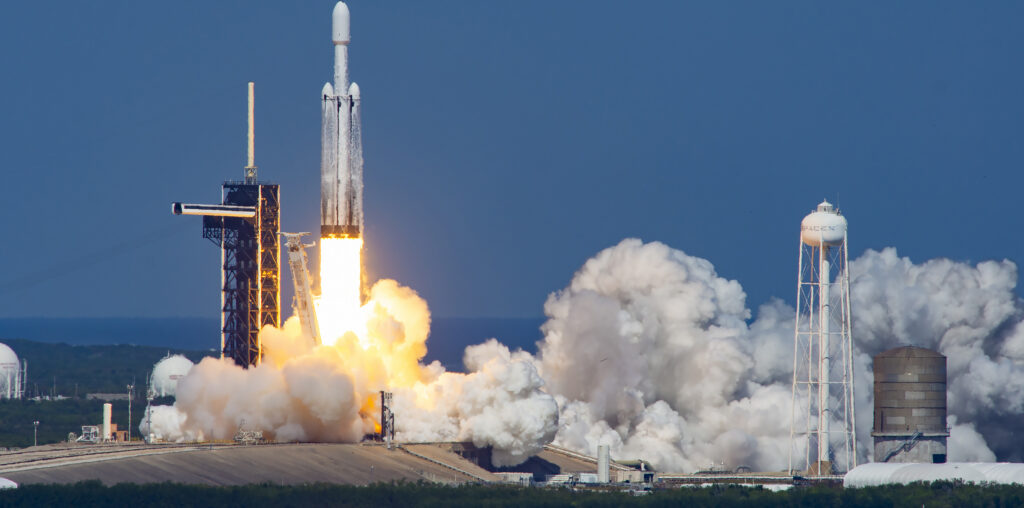 A white rocket lifts off from the launch pad, spewing billowing clouds of white vapor horizontally along the ground. A relatively short column of flames erupts from below the rocket just after liftoff. The early afternoon sky and the water in the foreground are similar, vibrant shades of blue.