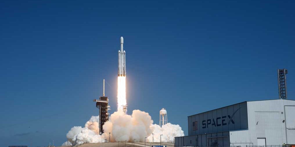 a rocket takes off with a plume of smoke underneath it and a space-x building to its right