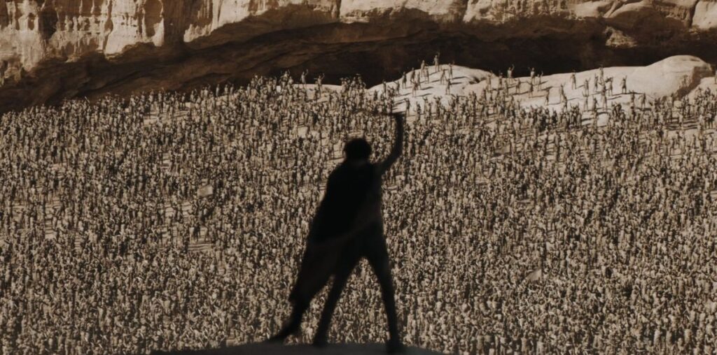 Image from the sci-fi movie Dune. Paul Atreides is holding a sword in the air as he is standing high above a crowd of thousands of people.