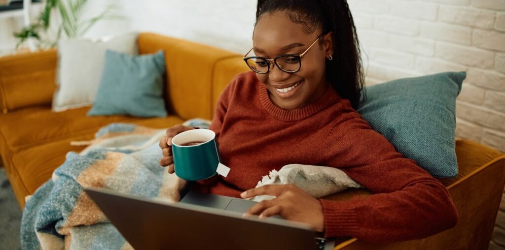 A woman sitting on a sofa with a throw, holding a cup of tea and using a laptop