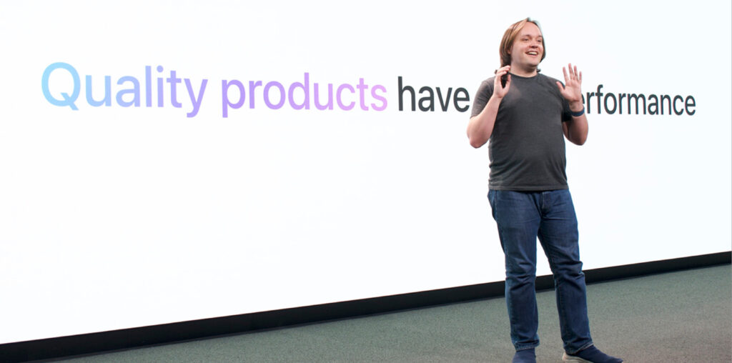 A person in a gray T-shirt and blue jeans stands on stage in front of a screen that reads "Quality products have great performance."