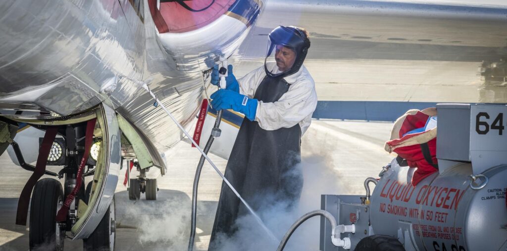 A man clad in a white protective suit, black apron, and blue gloves administers liquid oxygen from a tank to a parked aircraft via hose. Gas clouds hover near his feet and the man is wearing a helmet with a clear visor over his face. 