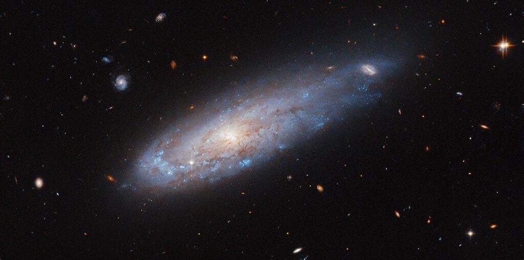 A spiral galaxy with a disk that glows visibly from the center. It has faint dust threaded through it. A spiral arm curves around the left edge of the disk and is noticeably denser with bright blue spots that hold hot and new stars. On the opposite side, the disk stretches out into a short tail where it covers a distant background galaxy. Other distant galaxies and some nearby stars are visible are also visible in this image.