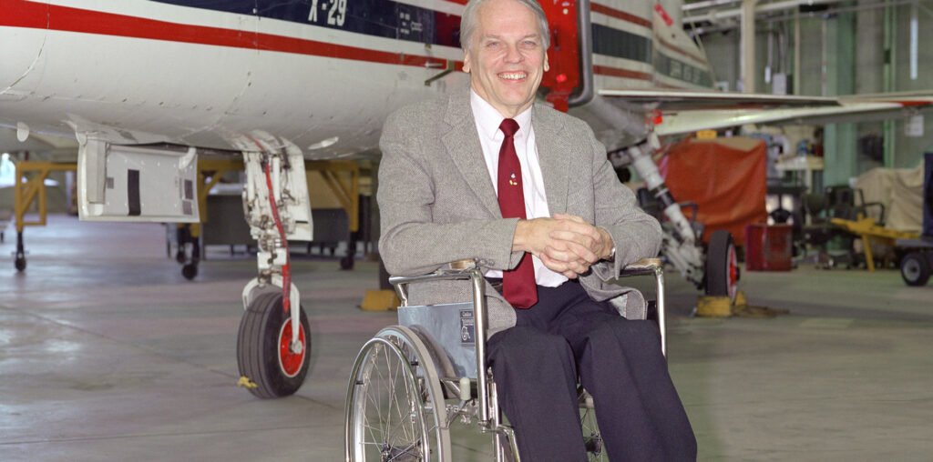 A man sits in a wheelchair with an experimental aircraft in the background.