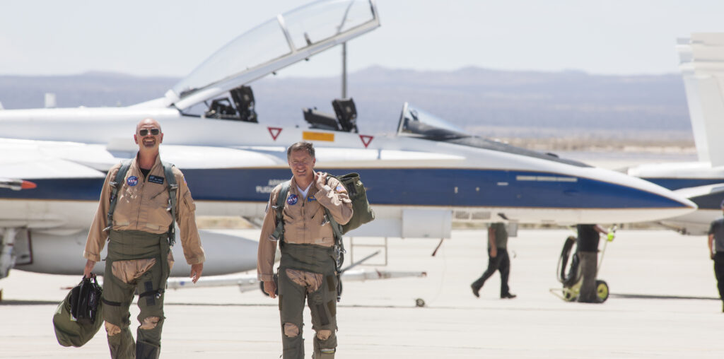 Two NASA test pilots walk toward the camera. Behind them is a blue and white F/A-18 research jet.