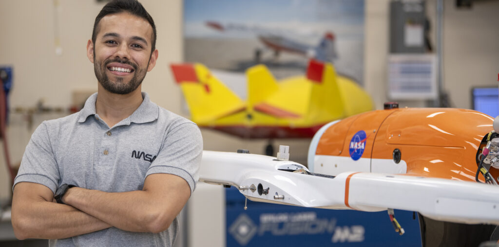 A man wearing a gray NASA shirt posing to the left of a subscale model or an aircraft that is orange and white with the NASA meatball on it. In the background, there are aviation posters located on the wall and another subscale model on display that is yellow and red.