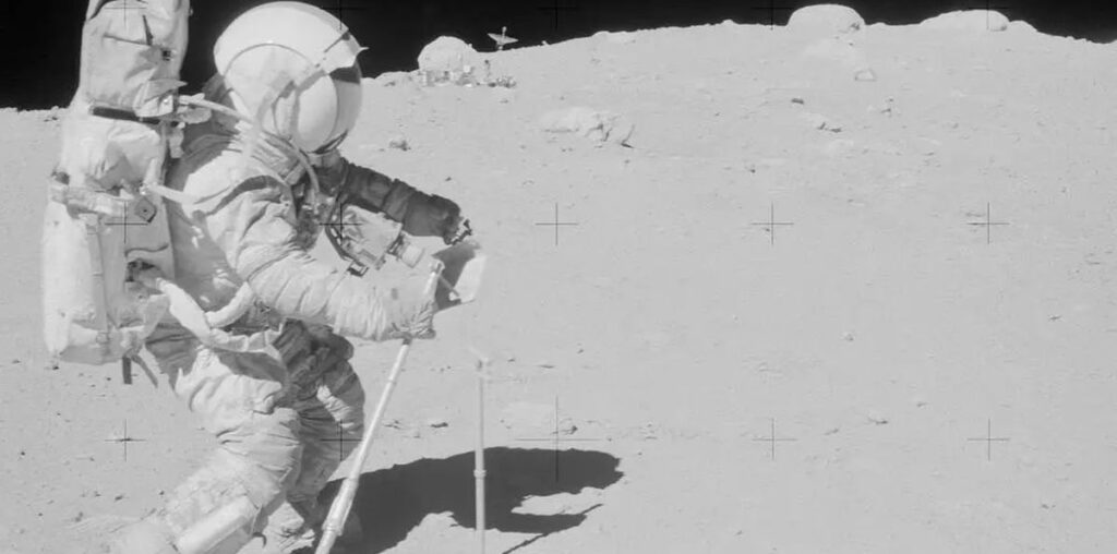 an astronaut in a bulky white suit walks on the dusty grey surface of the moon