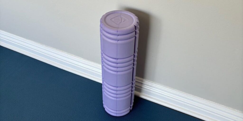 A close-up of the vertical purple foam roller on a blue yoga mat leaning against a beige wall.