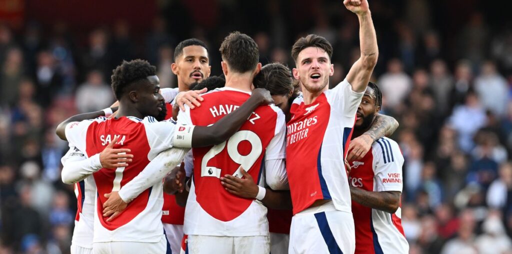 Arsenal players celebrate a goal in a huddle with Declan Rice turned to the camera pumping his fist.