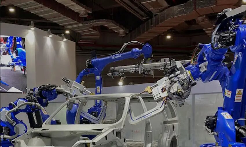 Shandong Golden Empire manufacturing demonstration with a white car frame in a conference hall and blue robotic arms on either side of the frame manipulating auto parts