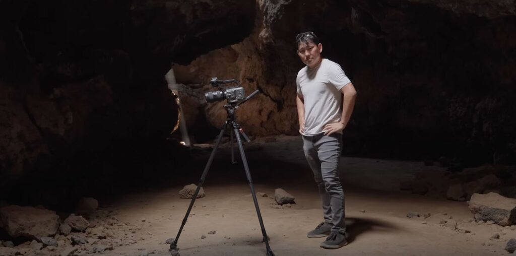 A person stands in a dimly lit cave with a video camera on a tripod. The person is wearing a light gray shirt, dark pants, and sunglasses on their head. The cave's rocky interior is visible in the background.