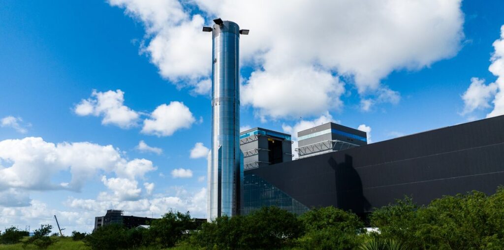 a tall silver rocket in front of a long low building and blue sky