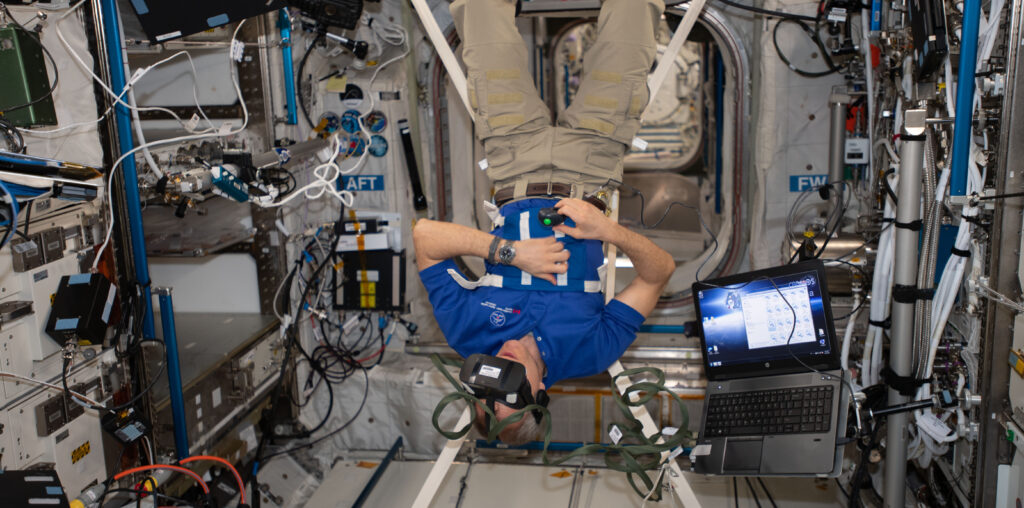 An astronaut floats upside down in the microgravity environment of the International Space Station, wearing a virtual reality headset.