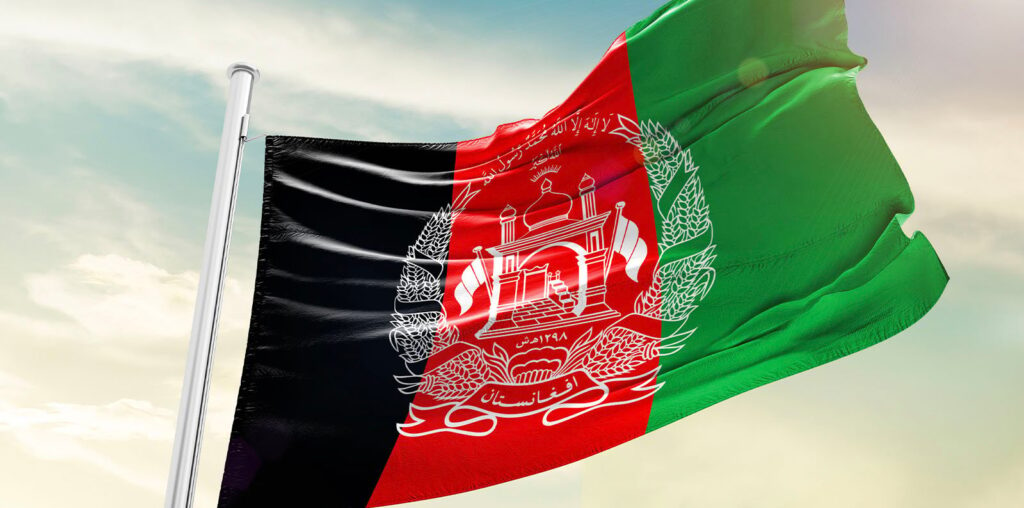 The flag of Afghanistan waves against a clear sky. It features vertical black, red, and green stripes with a white emblem in the center depicting a mosque with flags, sheaves of wheat, and Arabic script.