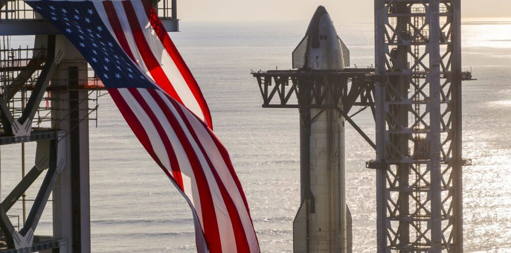 A giant American flag flutters in front of a silver SpaceX Starship rocket as it is hoisted atop its booster.