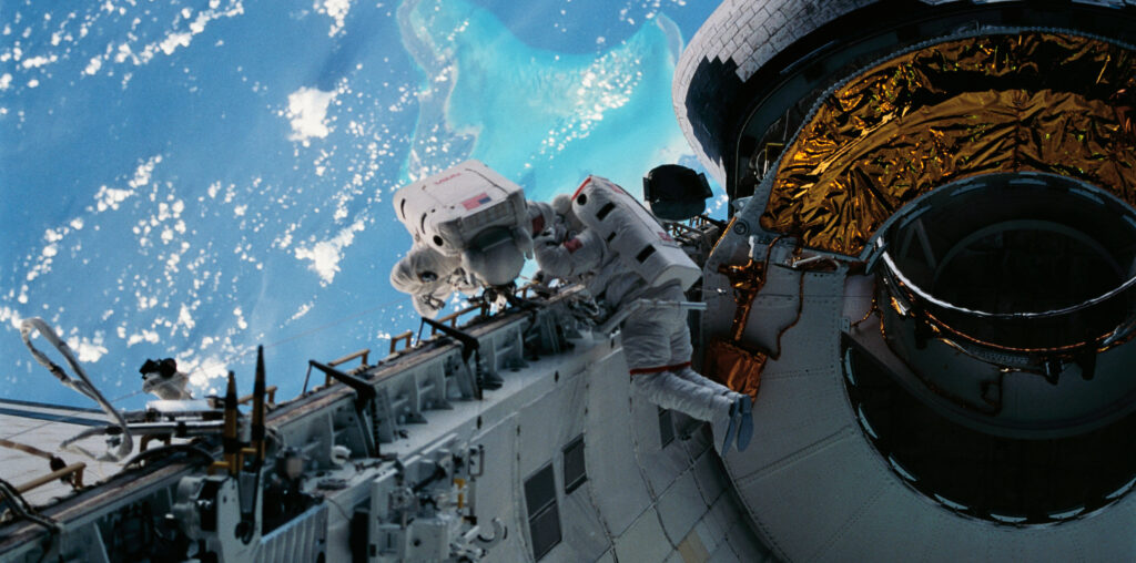 Two astronauts in white spacesuits inspect equipment outside of space shuttle Discovery during a spacewalk. The Caribbean Sea and part of the Bahama Islands chain is visible behind them.