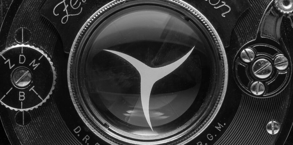 Close-up of an old camera lens with vintage markings. The black and white image highlights intricate details, including a prominent central icon resembling a metallic triangle. Surrounding the glass are mechanical dials and lettering, enhancing its classic look.