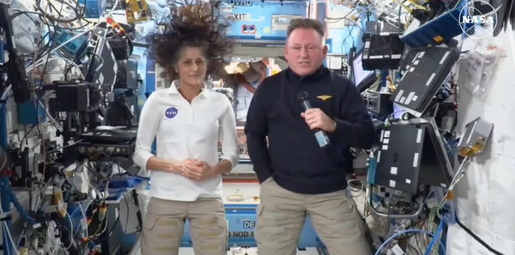 Two people floating in microgravity on the International Space Station. They are surrounded by lots of wires and other hardware.