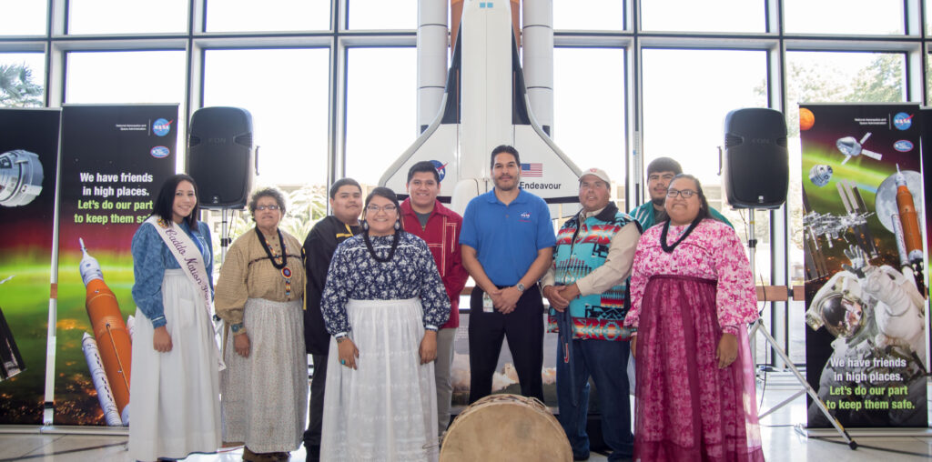 A group of Indigenous people and a NASA employee pose together in front of a model of the Space Shuttle Endeavour. The individuals wear traditional clothing, including sashes and beaded garments.