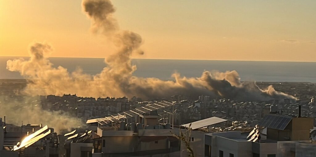 Smoke rises from buildings in a cityscape, illuminated by the setting sun. Rooftops with solar panels are visible in the foreground, and the sea stretches out on the horizon.