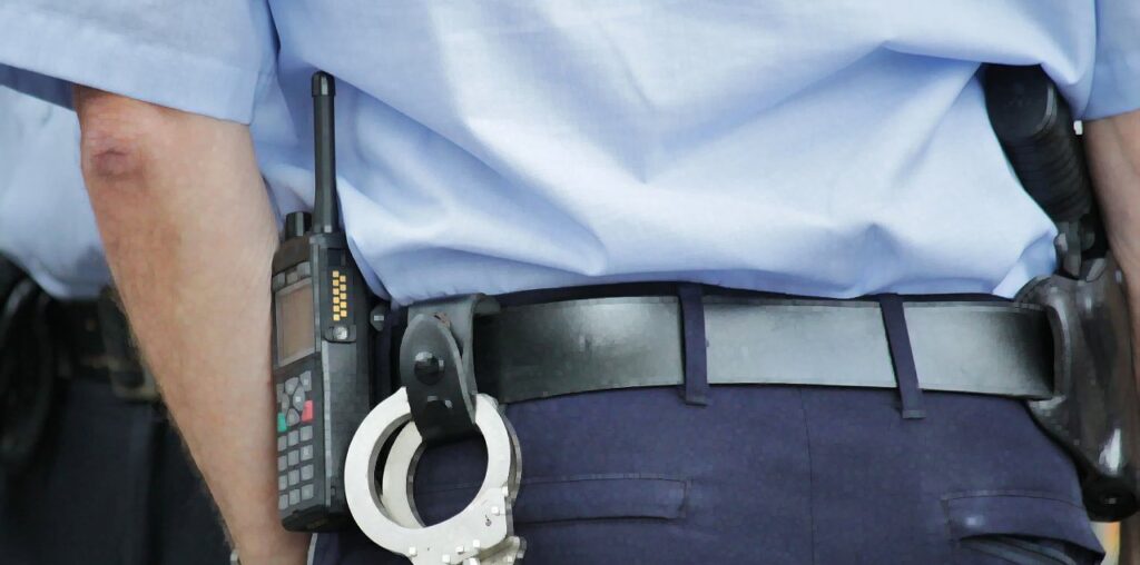 Close-up of a police-officer's belt from behind. We see a walkie-talkie, handcuffs, and a pistol in its holster.