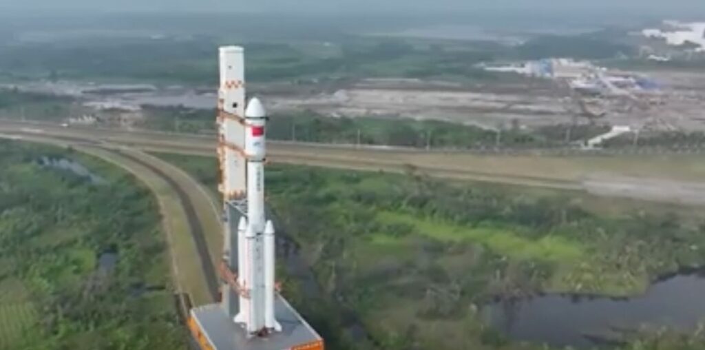 a white rocket rolls out to its launch pad on a track that passes greenery and a small lake