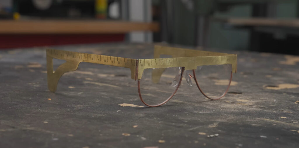 A set of brass safety glasses sit on a marred black workbench. The top and earpiece sections of the frames are in squarish brass plate and have ruler marks on them.