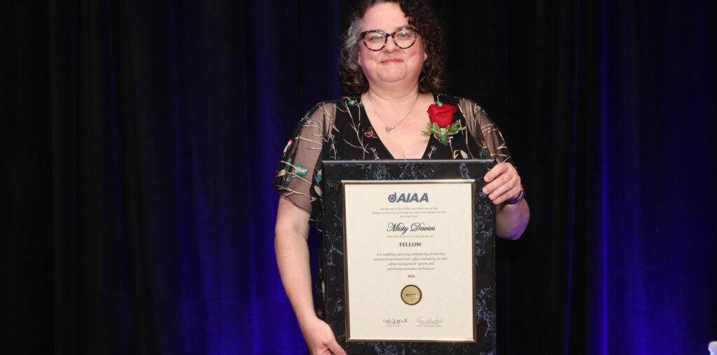 Misty Davies holding a framed AIAA Fellowship certificate with a red rose on her lapel, standing against a dark background.