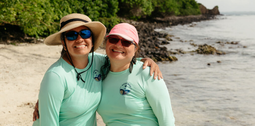 Two Puerto Rican women stand on a beach, both wearing teal longsleeve shirts and sunglasses. The one on the left has a tan, wide-brimmed hat, and the one on the right has a pink baseball cap.