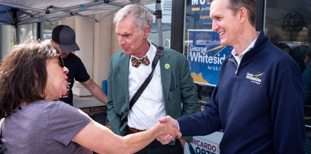 a smiling man in a blue sweater shakes hands with a woman while an older fellow in a bow tie looks on