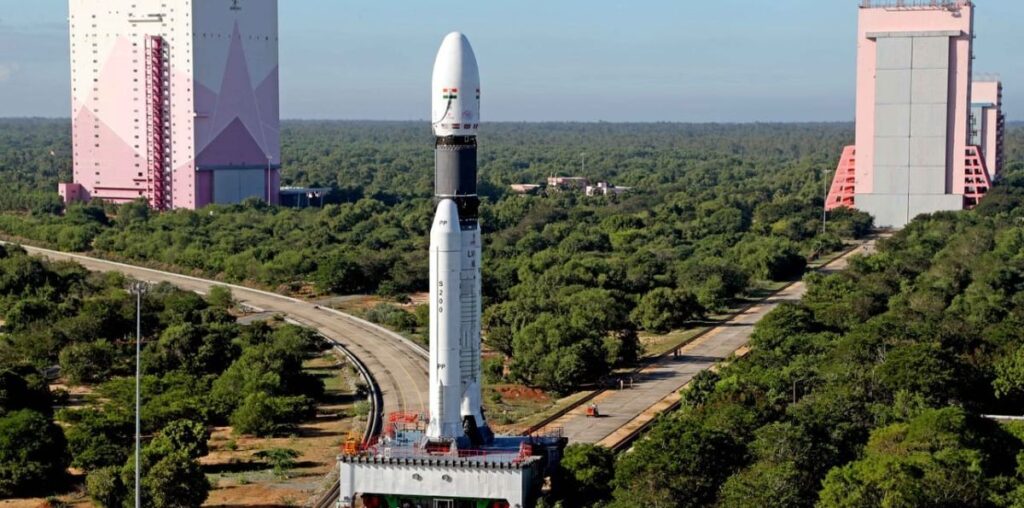 a white rocket with two white side boosters stands on a grey metal platform fixed on a track at the splitting toward two pink decorated hanger buildings