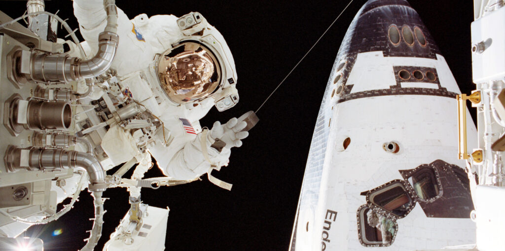 An astronaut in a white spacesuit appears on the left side of the image near a part. On the right in the back is part of the space shuttle Endeavour.