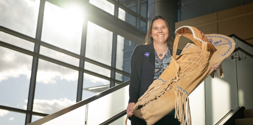 Tami, a woman of Native American heritage from the Delaware Nation, smiles standing on a staircase in a modern, sunlit building, holding a traditional handcrafted cradleboard adorned with intricate beadwork. She is wearing a paisley patterned shirt covered with a black sweater that has the NASA insignia on the right breast.