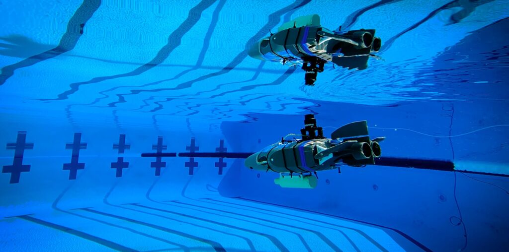 A small gray plastic robot shaped like a wedge (with propellers and flaps for steering) glides through the blue waters of a competition swimming pool.