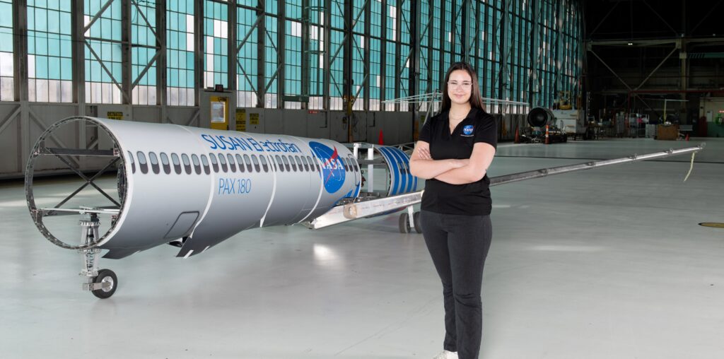 A woman wearing a black polo shirt with the NASA insignia, dark gray pants, and white shoes stands inside an aircraft hangar in front of a scaled aircraft model. The words “SUSAN Electrofan” and “PAX 180” are printed on a graphic along with fuselage, with a large NASA insignia in the center of the plane. The rear of the aircraft model, along with the wings and tail, are taken apart to display the inner components that make up its electrified propulsion system.