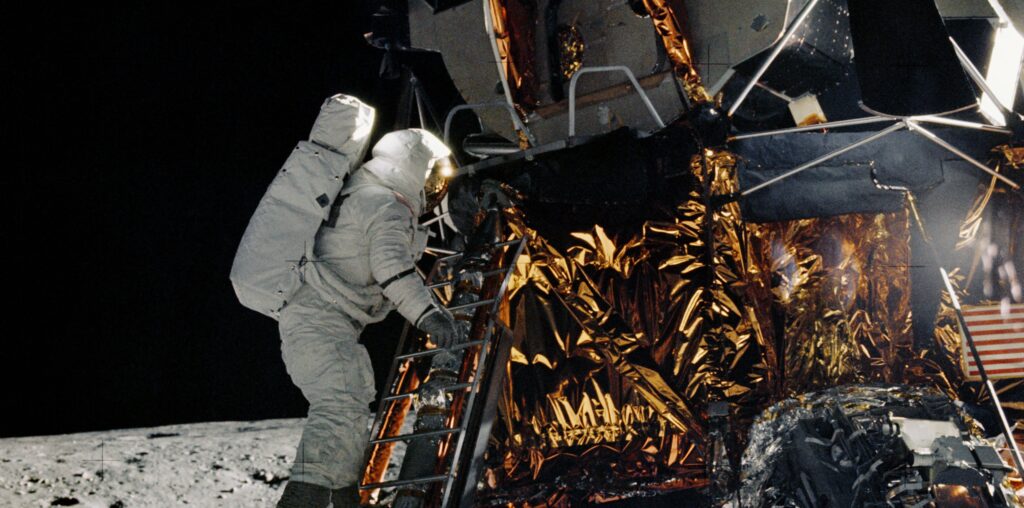 On the Moon, an astronaut in a white spacesuit climbs down a ladder on a lunar module. The lunar module is made of metal with gold and silver foil covering some of its lower half.