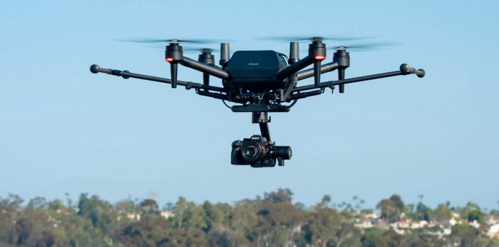 A quadcopter drone equipped with a camera hovers in the sky. The background shows a blurred landscape of trees and buildings under a clear blue sky.