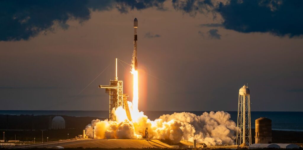 a black-and-white spacex falcon 9 rocket launches into a darkening evening sky.