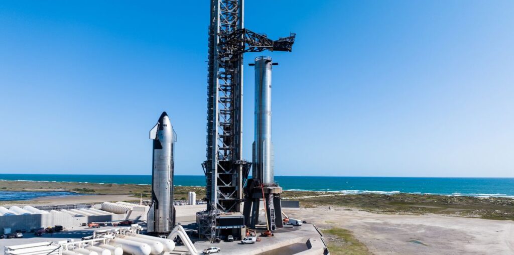 a silver rocket and silver spacecraft stand vertically next to a seaside launch tower