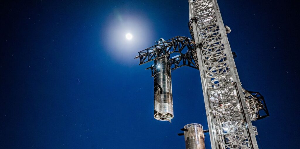 a large silver spacecraft is placed atop a booster by a launch tower at night