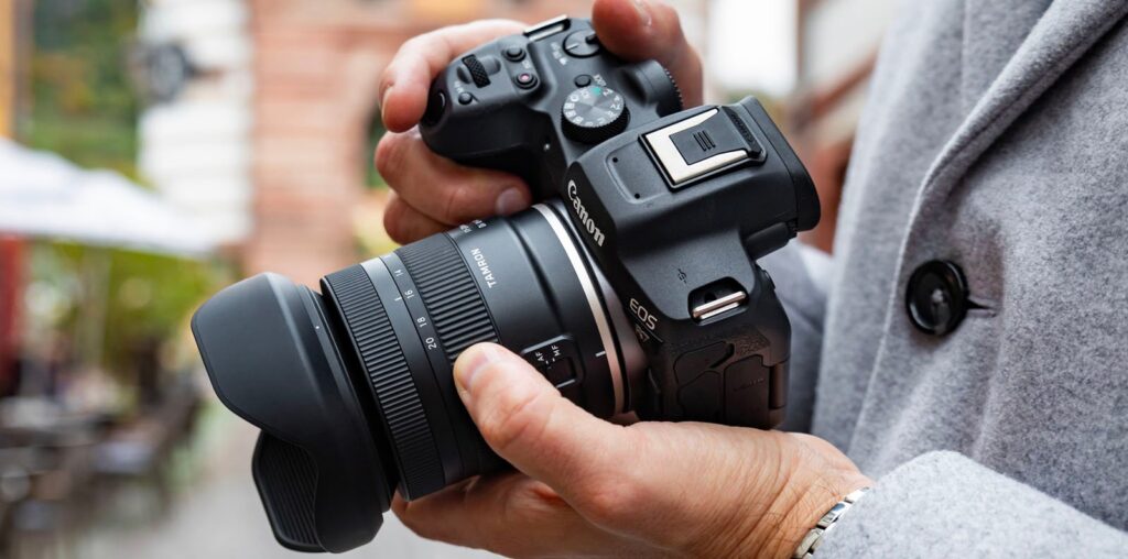 Person holding a Canon EOS camera with both hands, focusing on the camera lens. Background is slightly blurred, indicating an outdoor setting. Person is wearing a light gray coat.