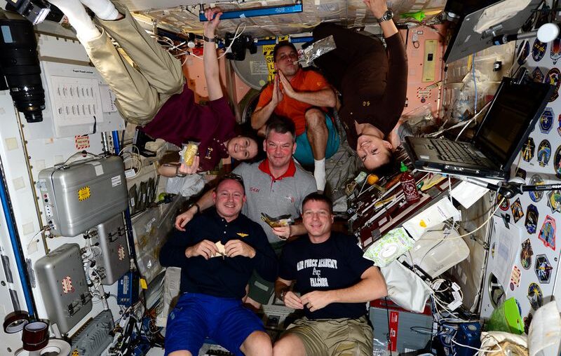 Three astronauts wearing black shirts, one astronaut wearing a burgundy shirt, one astronaut wearing a grey shirt with a red collar, and one astronaut wearing an orange shirt are all gathered together in a small space onboard NASA's International Space Station. Two astronauts (one in a burgundy shirt and one in a black shirt) are floating upside down. They are all eating.