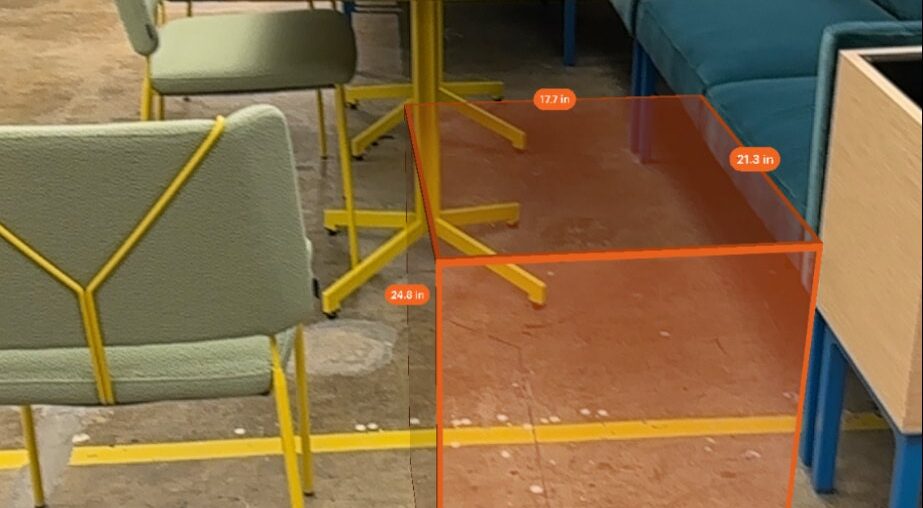 An orange rectangular prism placed in between furniture with measurements shown on each edge of the prism